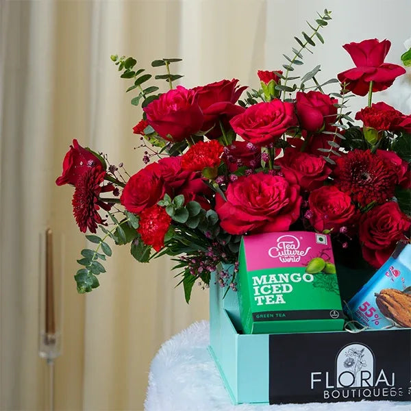 a vibrant mix of red carnations and red roses is arranged alongside a pack containing chocolate cookies and mango iced tea close up view
