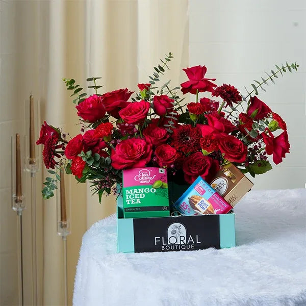 a vibrant mix of red carnations and red roses is arranged alongside a pack containing chocolate cookies and mango iced tea front view